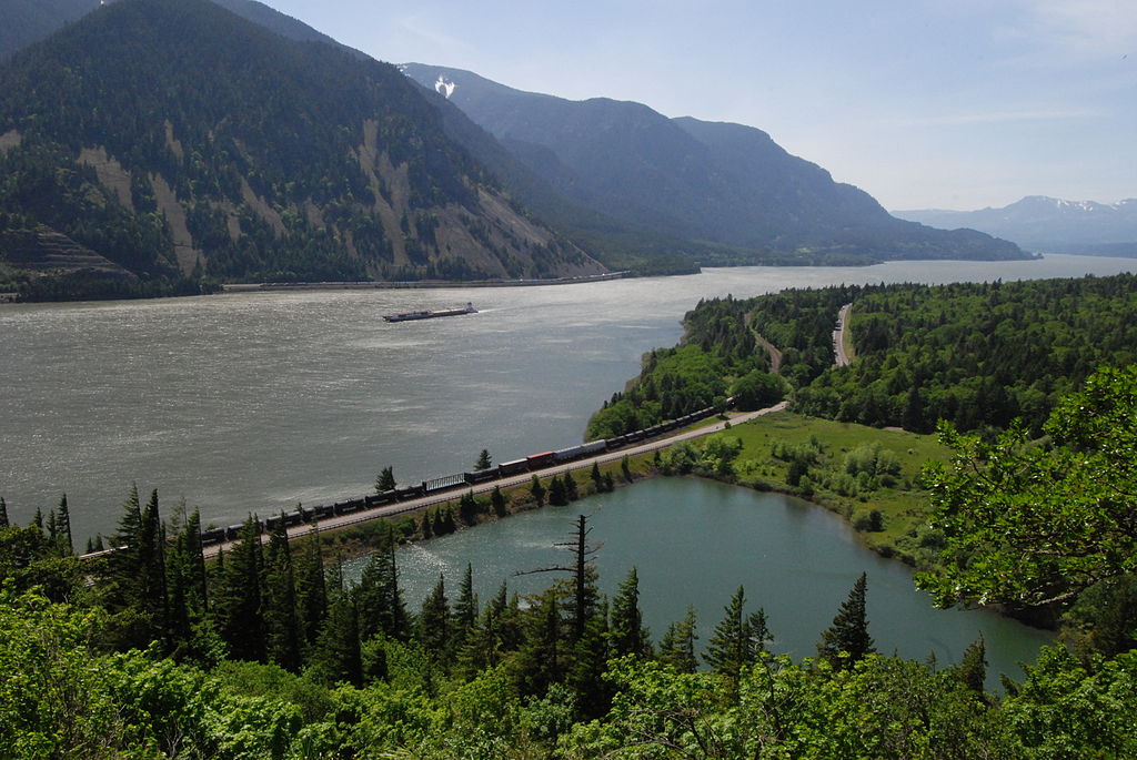 A picture of mountains and a river with a barge on the river and a railroad with a train on it in the foreground