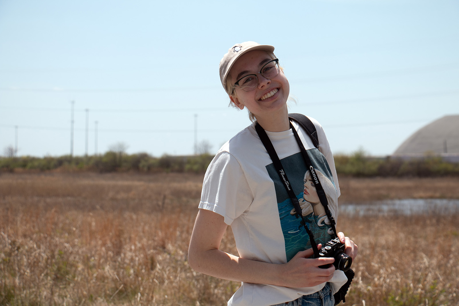 A young woman stands outside in a field while holding a camera that hangs from a neck strap she is wearing.