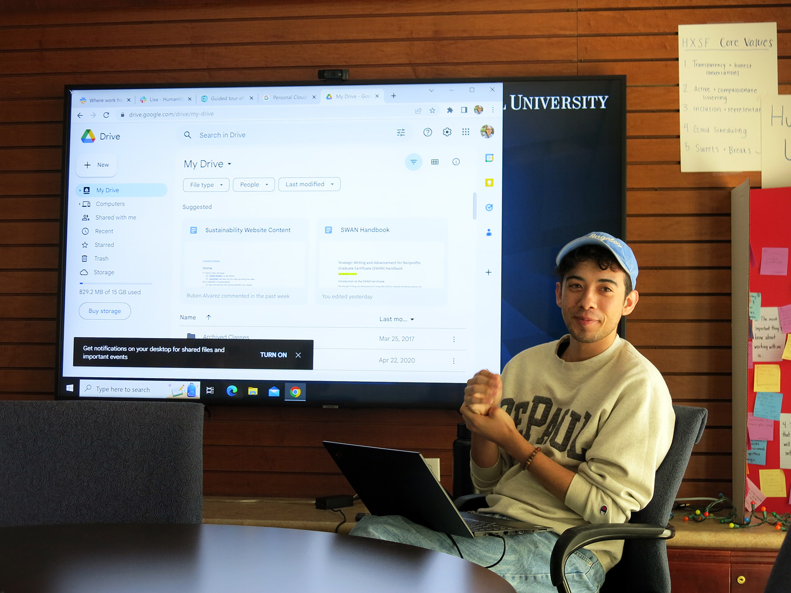 A young man sits in an office chair in front of a projected screen of a computer